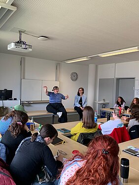 Teresa Reichl und Christine Ott sitzen auf einem Tisch in einem Kursraum. Viele Studierende sitzen im Raum und hören zu. Teresa Reichl gestikuliert mit ihren Armen.