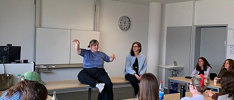 Teresa Reichl und Christine Ott sitzen auf einem Tisch in einem Kursraum. Viele Studierende sitzen im Raum und hören zu. Teresa Reichl gestikuliert mit ihren Armen.