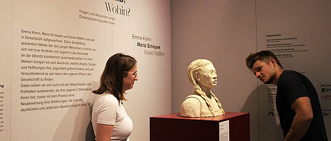 Zwei junge Menschen in einer Kunstausstellung. Sie betrachten eine Büste einer jungen Frau, die auf einem Sockel steht.