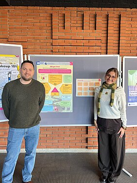 Die Studierenden Hannah Funke und Nicholas Cudd stehen im Gang des Philosophischen Gebäudes vor ihrem Präsentationsposter, das an eine Stellwand gehängt ist.