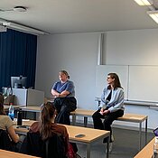 Teresa Reichl und Christine Ott sitzen auf einem Tisch in einem Kursraum und sprechen. Die Studierenden hören zu.