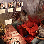 Zwei junge Menschen, die auf einem Sitzsack sitzen. Sie tragen Kopfhörer und hören sich Audiodateien an.