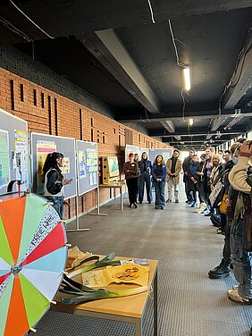 Eine Studentin steht im Gang des Phil-Gebäudes vor einer langen Stellwand mit verschiedenen Poster. Sie hält einen Vortrag über ihr Projekt. Ihr gegenüber steht das Publikum bestehend aus ca. 20 Studierenden und Mitarbeitenden der Uni. Im Vordergrund des Fotos sieht man das Glücksrad.