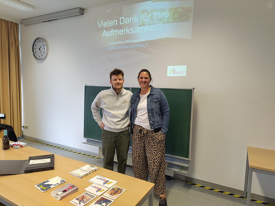 Foto von Elena Riedel und Florian Bötsch. Sie stehen im Raum, in dem das Mittagsgespräch stattgefunden hat. Vor ihnen auf dem Tisch liegen einige Exemplare von Publikationen des Verlags Rotscheibe.