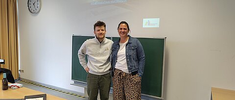 Foto von Elena Riedel und Florian Bötsch. Sie stehen im Raum, in dem das Mittagsgespräch stattgefunden hat. Vor ihnen auf dem Tisch liegen einige Exemplare von Publikationen des Verlags Rotscheibe.