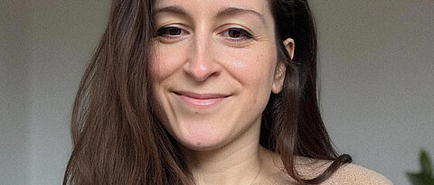 Porträtfoto von Anne Sauer. Person mit langen dunkelbraunen Haaren und hellbraunem Pulli lächelt in Kamera.