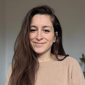 Porträtfoto von Anne Sauer. Person mit langen dunkelbraunen Haaren und hellbraunem Pulli lächelt in Kamera.